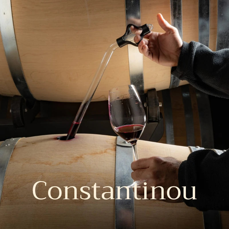 Person pouring red wine from a decanter into a glass with a wooden barrel in the background, featuring the brand 'Constantinou'.